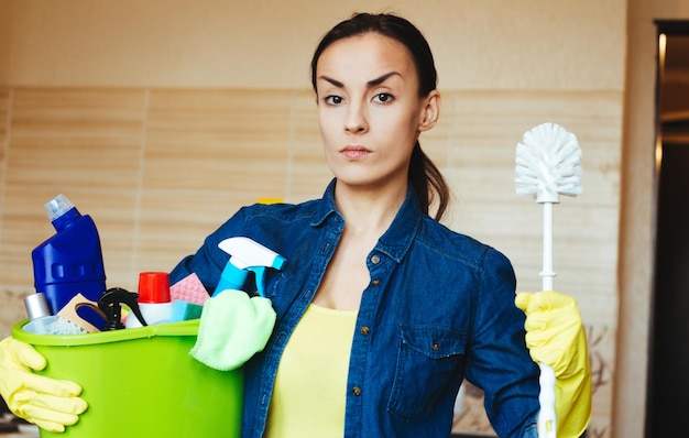 Beautiful woman in protective gloves is holding a bucket with things for cleaning looking at camera and smiling before start of house cleaning