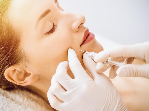 Beautiful woman receiving beauty injections with closed eyes in sunny medical center. Beautician doctor hands doing beauty procedure to female face with syringe. Cosmetic medicine and surgery concept.