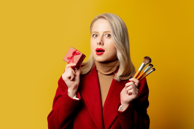 Beautiful woman in red coat with brushes and gift box on yellow wall