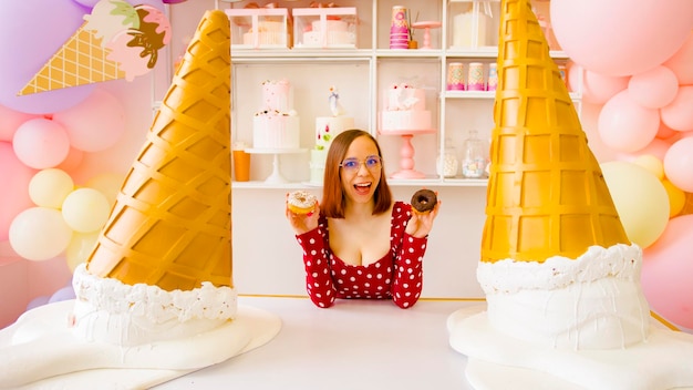 A beautiful woman in a red dress and glasses holds a white donut in one hand and a chocolate donut in the other hand and showing them to the camera in a cafe