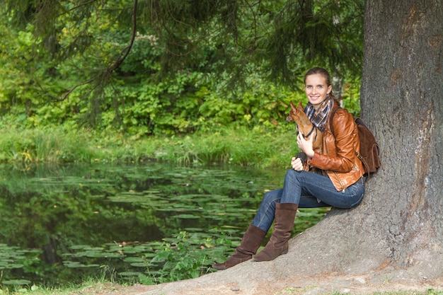 The beautiful woman resting outdoors and sitting at a tree with a little dog on hands on background of nature.