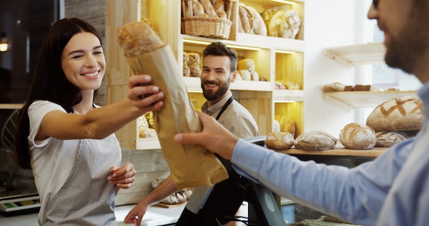 Beautiful woman, seller in the bakery shop, selling baguettes to the man while her male handsome co-worker helping her. At the counter. Indoor