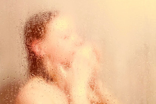 Beautiful woman in the shower behind glass with drops warm glow