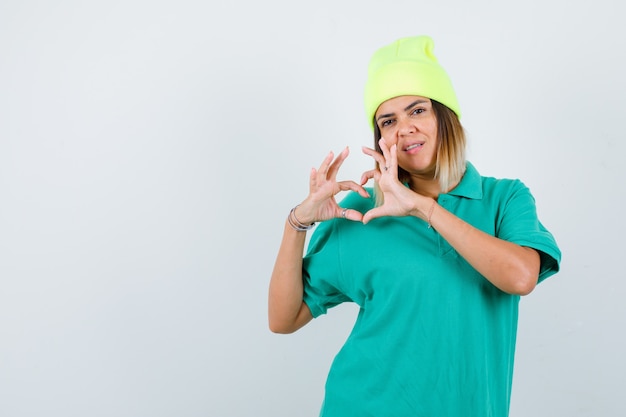 Beautiful woman showing heart gesture in polo t-shirt, beanie and looking confident. front view.