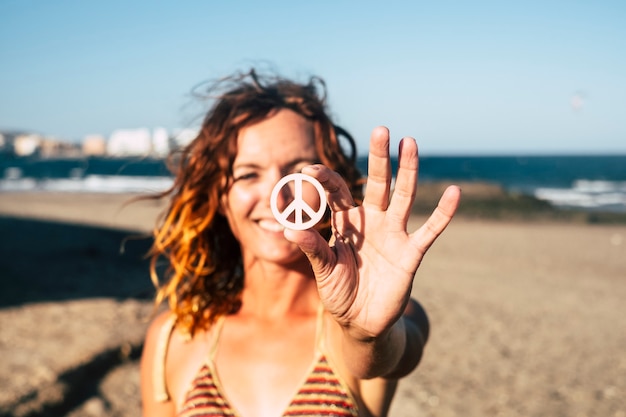 Beautiful woman showing the symbol of the peace at the beach alone and isolated with the sea and the sand of the beach at the background - caucasian girl smiling and looking at the camera