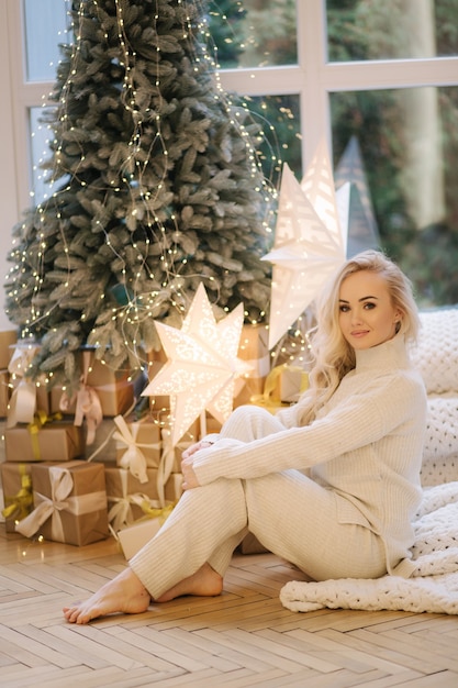 Beautiful woman sitting on windowsill by Christmas tree. Blond hair woman in white costume have rest