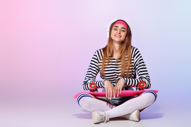 Beautiful woman sitting with skateboard and listening to music on pink background