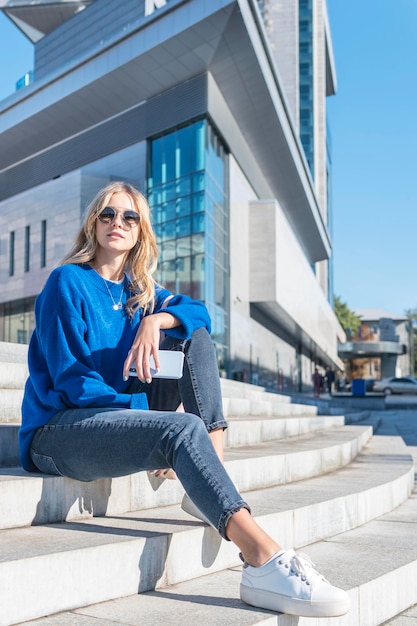 beautiful woman in sunglasses sits on the steps of a large modern office building.