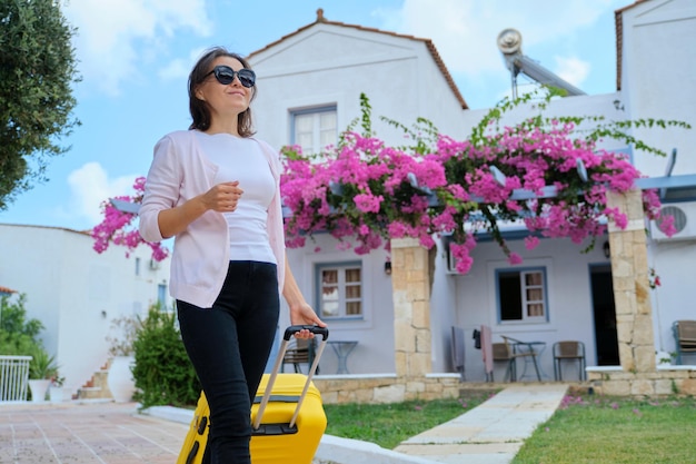 Beautiful woman tourist walking with suitcase on territory of resort spa hotel. White blue hotel houses, blooming gardening with pink flowers background. Travelling, vacation, leisure, weekend, people