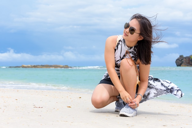 Beautiful woman tourist wearing sunglasse squatting binding rope sneaker on the beach near the sea during the stroll on Tarutao island in the summer vacation, Satun, Thailand