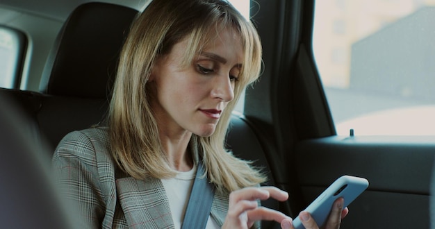 Beautiful woman typing on her smartphone in the car Lady manager looking at phone in the back seat of a car