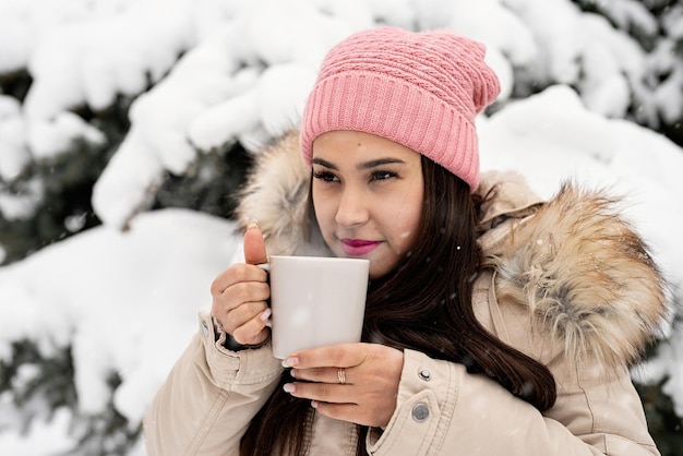 Beautiful woman in warm winter clothes holding cup drinking hot tea or coffee outdoors in snowy day