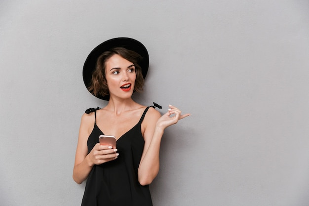 beautiful woman wearing black dress and hat smiling while holding smartphone, isolated over gray wall