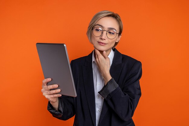 Beautiful woman wearing black official style suit looking at tablet isolated over orange background