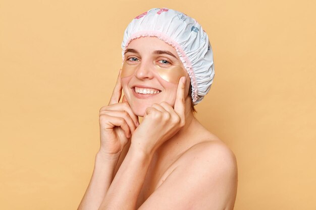 Beautiful woman wearing shower cap standing isolated over beige background posing with patches taking care of her under eye skin touching her hydrogel patches