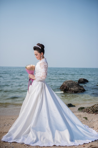 Beautiful woman in wedding dress holding flower standing on a beach.