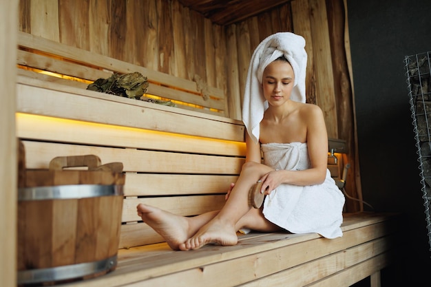 Beautiful woman in white bathrobe and towel on head doing body massage with dry wooden brush