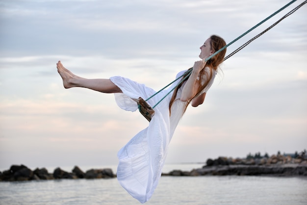 Beautiful woman in white outfit rides a rope swing over water. Hen party at the sea.