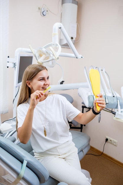 A beautiful woman in a white T-shirt sits in a dental office, looks in the mirror, smiles and holds a toothbrush in her hands. Girl with braces shows oral care. Dentistry, dental treatment