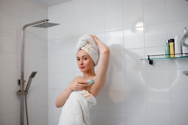 Beautiful woman in white towel shaving her armpits with razor in shower cabin Depilatory procedure at bathroom