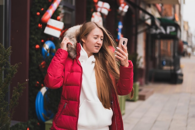 Beautiful woman in winter clothes walks down the street decorated with New Year's decor and communicates by phone