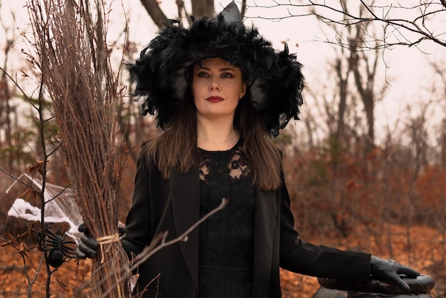 Beautiful woman in witches hat and costume holding broom near the cauldron in autumn forest Halloween concept Selective focus