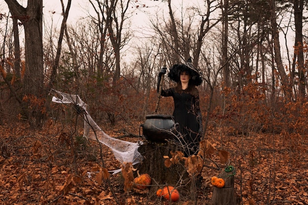 Beautiful woman in witches hat and costume preparing a potion in cauldron in autumn forest Halloween concept Selective focus