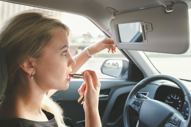 Beautiful woman with blond hair driving a car paints her lips with lipstick