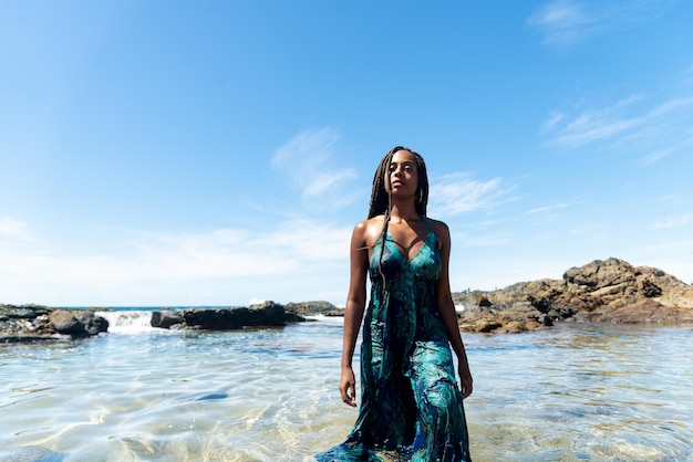 Beautiful woman with braids and blue dress in the beach water walking towards the camera