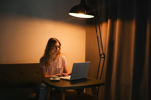 Beautiful woman with cup of coffee working on laptop in evening Girl with notebook in dark room Di