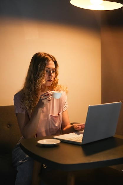 Beautiful woman with cup of coffee working on laptop in evening Girl with notebook in dark room Di