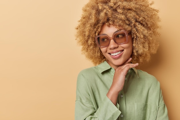 Beautiful woman with curly bushy hair touches jawline gently smiles gladfully wears sunglasses and shirt notices something pleasant aside poses against brown background blank space for text