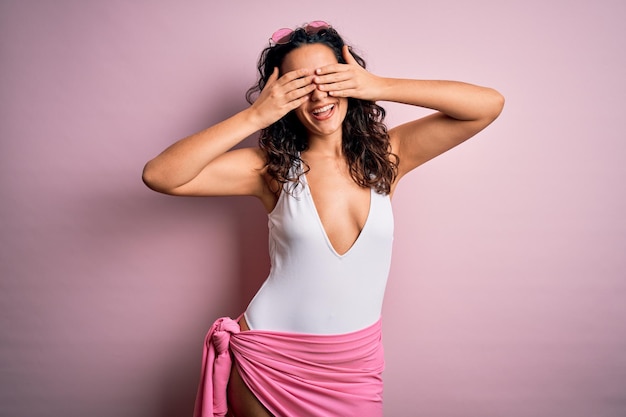 Beautiful woman with curly hair on vacation wearing white swimsuit over pink background covering eyes with hands smiling cheerful and funny Blind concept