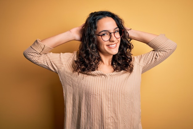 Beautiful woman with curly hair wearing striped shirt and glasses over yellow background relaxing and stretching arms and hands behind head and neck smiling happy