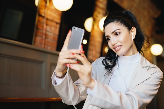 Beautiful woman with dark hair tied in pony tail making selfie while sitting in cafe using her smartphone