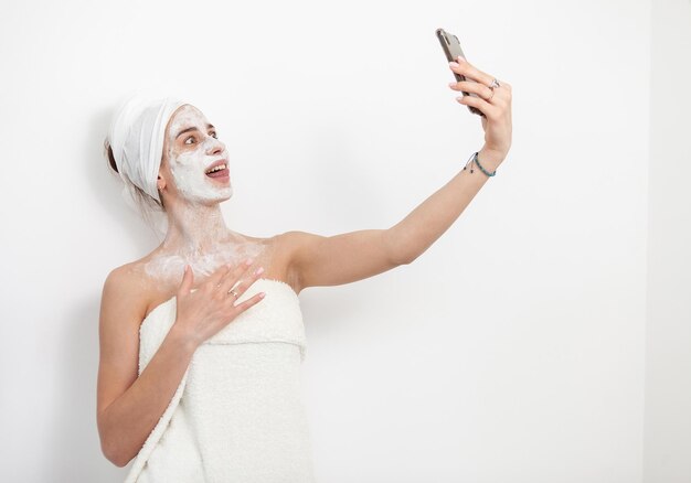 Beautiful woman with facial mask makes selfie on smartphone on white background Skin care concept