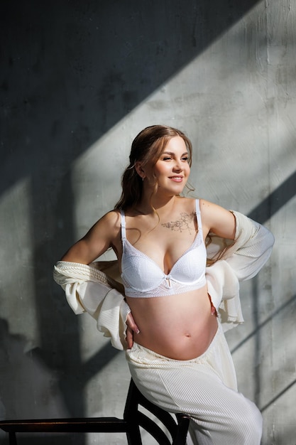 A beautiful woman with fair hair is pregnant in a white elegant suit sitting on a chair on a gray background Fashion for pregnant women A stylish woman is pregnant