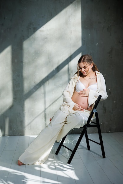 A beautiful woman with fair hair is pregnant in a white elegant suit sitting on a chair on a gray background Fashion for pregnant women A stylish woman is pregnant