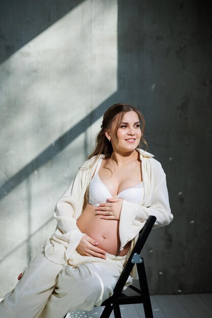 A beautiful woman with fair hair is pregnant in a white elegant suit sitting on a chair on a gray background Fashion for pregnant women A stylish woman is pregnant