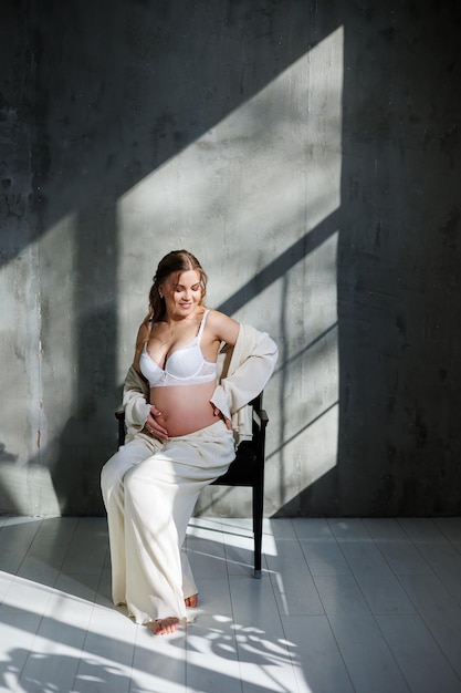 A beautiful woman with fair hair is pregnant in a white elegant suit sitting on a chair on a gray background Fashion for pregnant women A stylish woman is pregnant