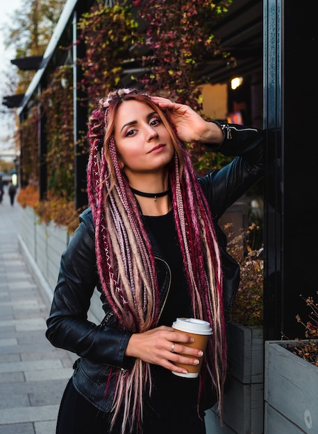 Beautiful woman with long pink dreadlocks in black clothes on the street holds a cup of coffee