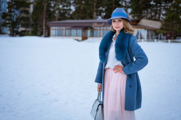 Beautiful woman with makeup, in a blue coat and hat, posing in nature on the snow