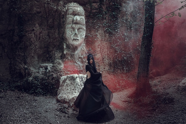 A beautiful woman with pale skin in a black dress and in the black crown. Gothic look. Outfit for halloween. A woman with colored smoke against the sculpture of an idol