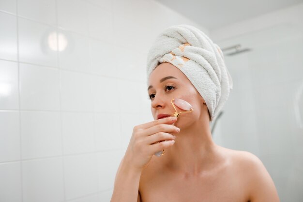 Beautiful woman with perfect skin wearing white towel after shower making face massage using a jade face roller with natural quartz stone in bathroom Natural cosmetics concept wrinkle smoothing