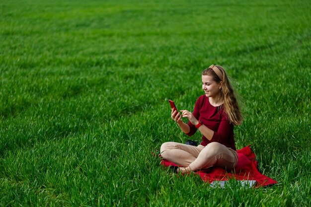 Beautiful woman with a phone in her hands sits on the grass. The girl looks at herself in the camera of the phone and takes selfie photos. She smiles and enjoys a warm day. Concept photo on smartphone