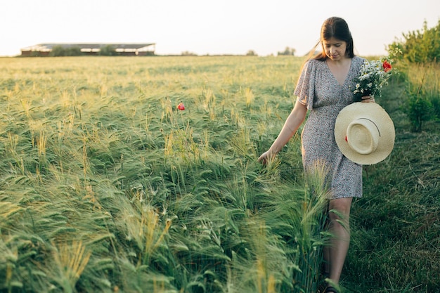 Beautiful woman with straw hat and wildflowers enjoying sunset in barley field Atmospheric tranquil moment slow life Stylish female gathering flowers and walking in evening summer countryside