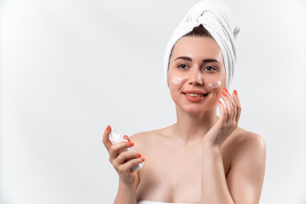 Beautiful woman with a towel on her head smiles and smears face cream on a white background isolated