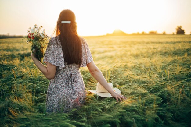 Beautiful woman with wildflowers enjoying sunset in barley field Atmospheric tranquil moment rustic slow life Stylish female gathering flowers and relaxing in evening summer countryside