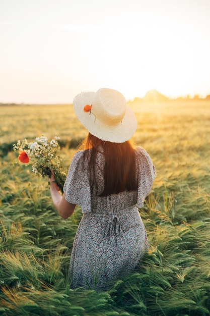 Beautiful woman with wildflowers enjoying sunset in barley field Atmospheric tranquil moment rustic slow life Stylish female gathering flowers and relaxing in evening summer countryside