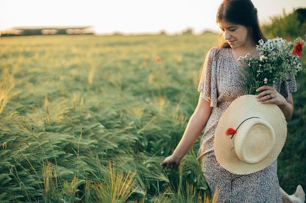 Beautiful woman with wildflowers and straw hat walking in barley field in sunset light Stylish female relaxing in evening summer countryside and gathering flowers Atmospheric tranquil moment